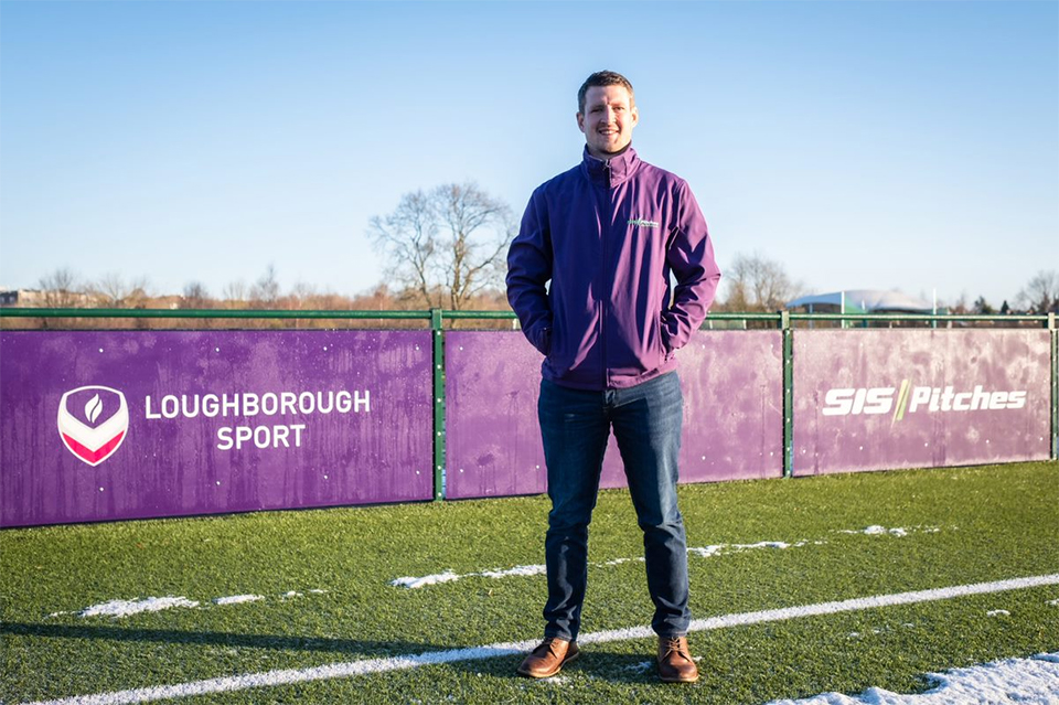 Joe stands on a pitch in front of purple signs. The signs have the Loughborough Sport and SIS Pitches logos on them. The image is taken on a clear day in January. There are some trees in the background.