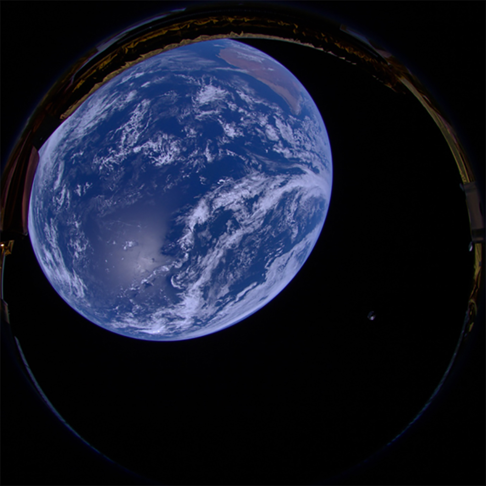 Photo of Earth from Low Earth Orbit using onboard camera on the HAKUTO-R Lunar Lander Spacecraft. There is an image of earth taken from space. The planet’s blue colour glows against the darkness. A small grey shape is on the bottom right of the image - this is the SpaceX Falcon rocket in its second stage.