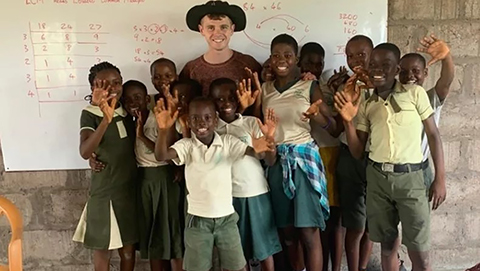 Chris Jones posing in a classroom in front of a whiteboard with red pen writing on it with a group of kids.