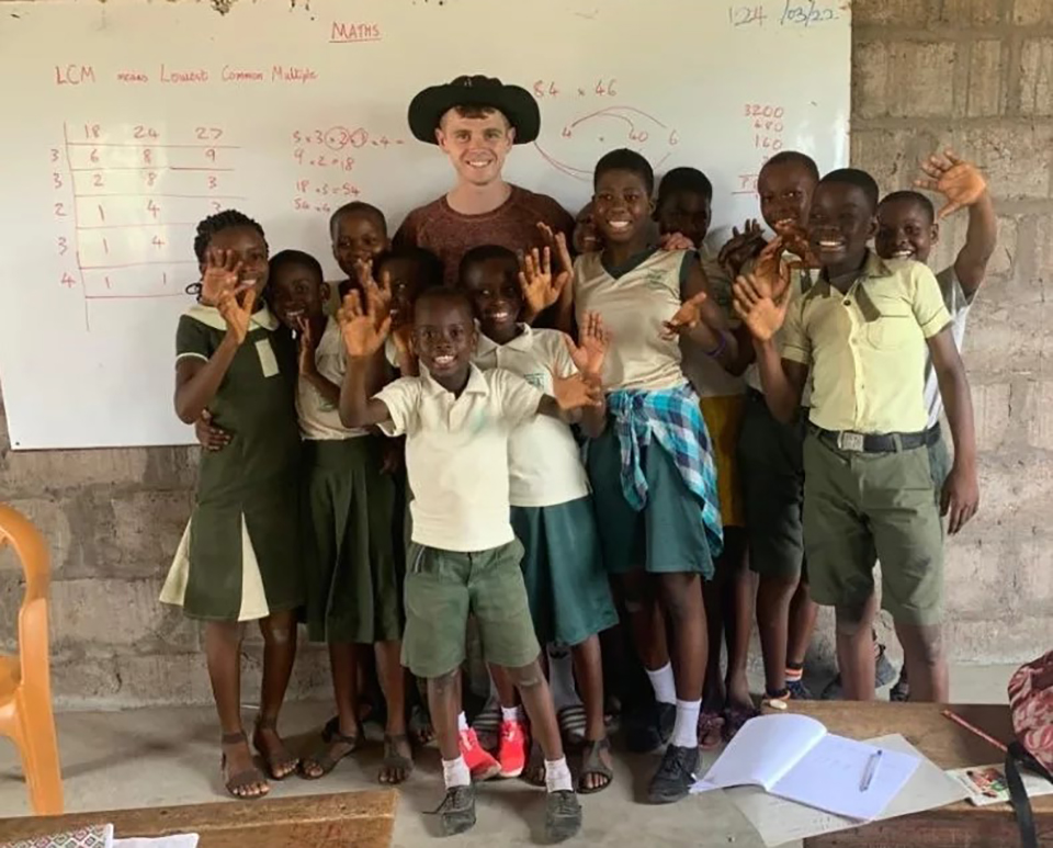 Chris Jones posing in a classroom in front of a whiteboard with red pen writing on it with a group of kids.