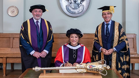 Deborah Cadman seated in the centre wearing a red robe and hat. To the left is Vice-Chancellor Professor Nick Jennings and on the right is Chancellor Lord Seb Coe. They both wear graduation robes.