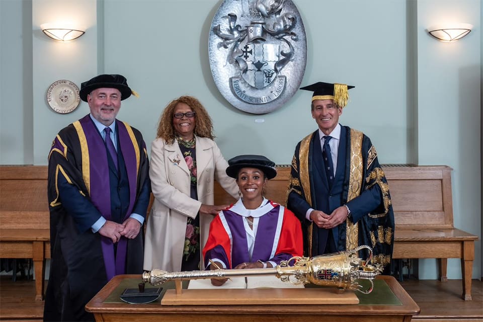 Ebony-Jewel Rainford-Brent is seated at a desk wearing a red robe and hat. Standing to the left is Vice-Chancellor Professor Nick Jennings and her mother, Janet Rainford. On the right is Chancellor, Lord Sebastian Coe. 