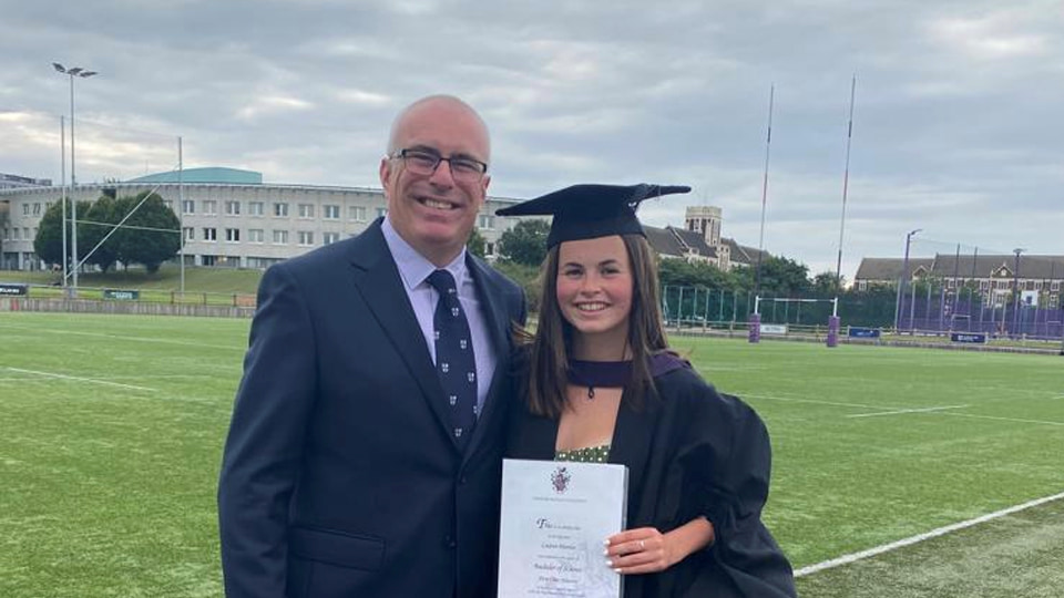 Chris and Lauren Martin stand together on the rugby pitch. Lauren wears a cap and gown and is holding her degree certificate. Chris wears a suit and a Loughborough tie. In the background there’s a rugby post, the business school building, and the Hazlerigg and Rutland buildings. 