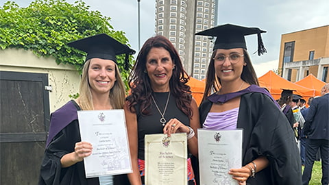 Caitlin, Caroline and Shania Robba each holding their degree certificates. Caitlin and Shania are wearing graduation caps and gowns. The trio stand on grass and in the background is Towers Hall.