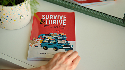 An image of the Survive & Thrive book on a desk in front of a mirror. It is next to a plant pot and someone’s hand is opening the front cover of the book, which is red with an illustration of a loaded car on the front.