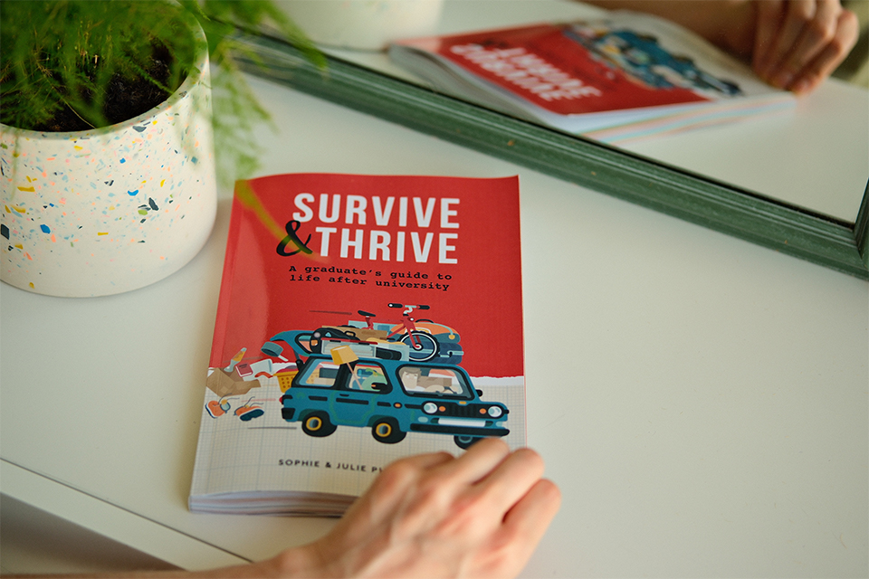 An image of the Survive & Thrive book on a desk in front of a mirror. It is next to a plant pot and someone’s hand is opening the front cover of the book, which is red with an illustration of a loaded car on the front.