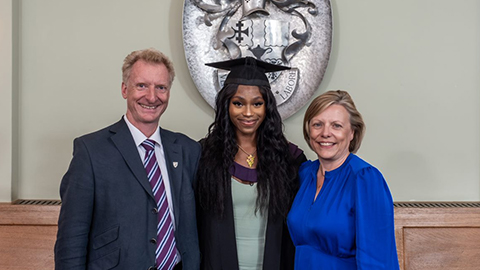 Tobi Omiyale stands in the centre of the image wearing a graduation cap and gown. Steven is on the left wearing a suit and Sally-Ann is on the right of the image wearing a blue dress. They all stand together in the Billiard Room in the Hazlerigg Building in front of wooden benches. There is a silver-colour university crest on the wall. 