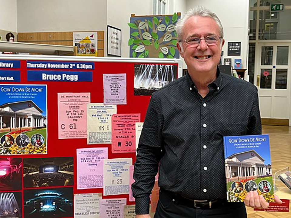 Bruce Pegg posing with his book at the Central Library in front of a decorated stand.