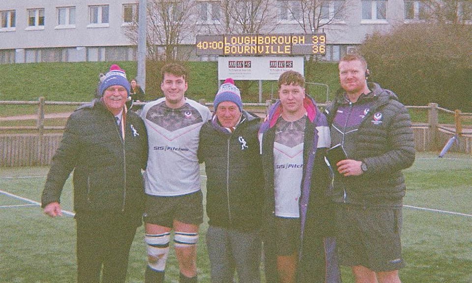Five people stand in front of a rugby scoreboard on the Loughborough campus. The image features Kevin Lucyszyn, Rex Hazeldine, student rugby players and a rugby coach.