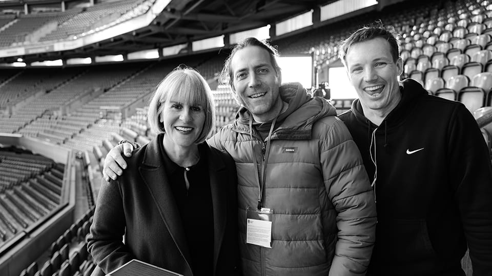 Sue Anstiss with Director of Photography Ben Marlow and alumnus Jack Tompkins, Director and Producer. The trio stand together inside a stadium.