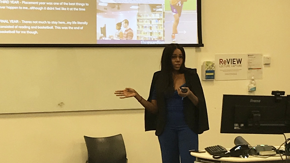 A photo of Aisha standing at the front of the lecture room. There is a large screen and computer also in the image.