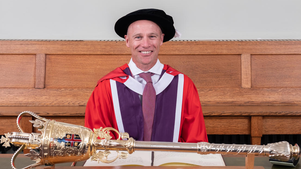 Danny is pictured wearing a red graduation gown and cap. He is seated and the University Mace is on the table in front of him. Danny received his Honorary Degree from Loughborough in 2019.