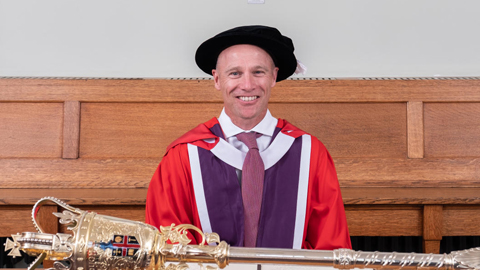 Danny is pictured wearing a red graduation gown and cap. He is seated and the University Mace is on the table in front of him. Danny received his Honorary Degree from Loughborough in 2019.