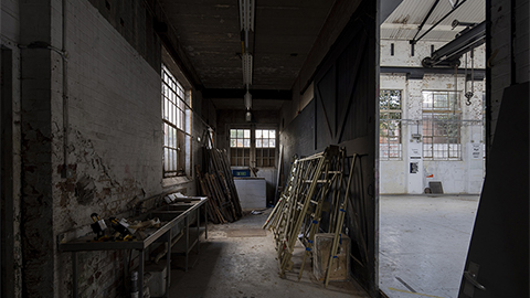 Inside the Generator building. The image is of a corridor with a door at the end. There are old tables and pieces of wood leaned against the walls. To the right of the image is a view of the next room, which is lighter, with windows in view.