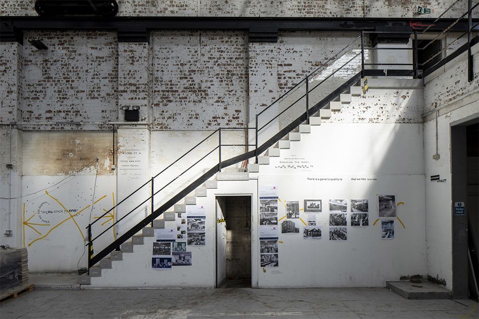 An image of the inside of the Generator building. There is a brick wall, and a side image of a set of stairs. There are a few photographs on the wall. The wall is painted white and is distressed on the brickwork. 