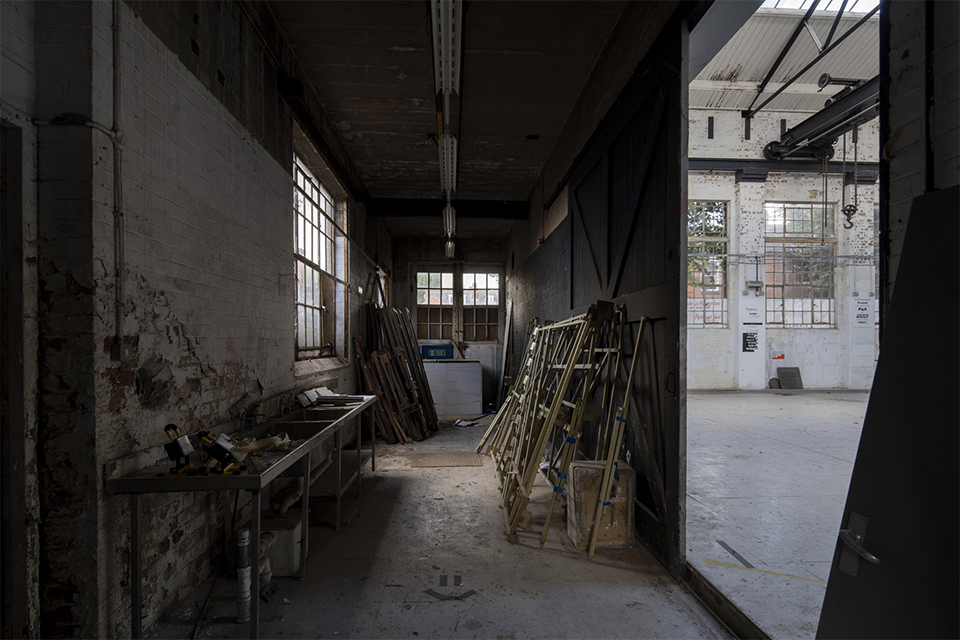 Inside the Generator building. The image is of a corridor with a door at the end. There are old tables and pieces of wood leaned against the walls. To the right of the image is a view of the next room, which is lighter, with windows in view.