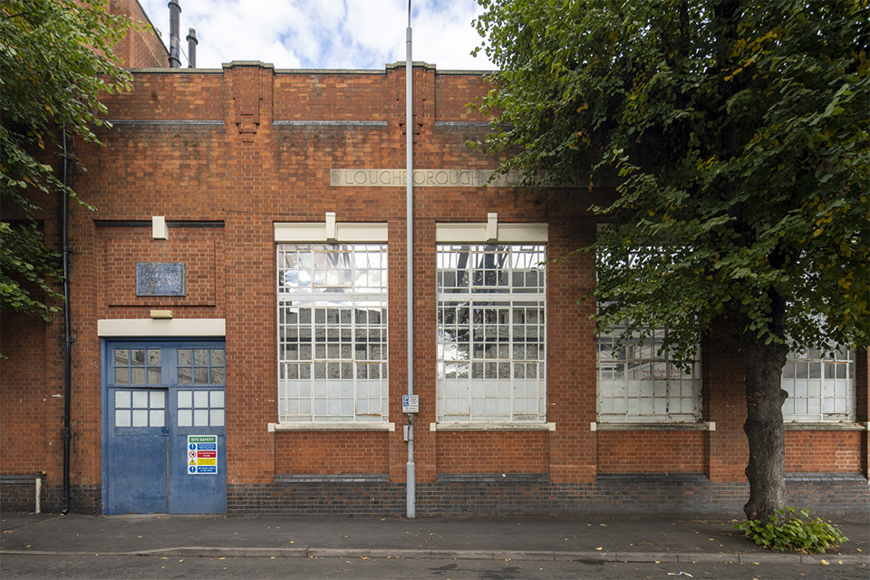 The outside of The Generator Building. It is a brown brick building with a blue door and large windows. There are also a couple of trees. There is a plaque on the side of the building for Loughborough College. Only the word “Loughborough” is in shot.