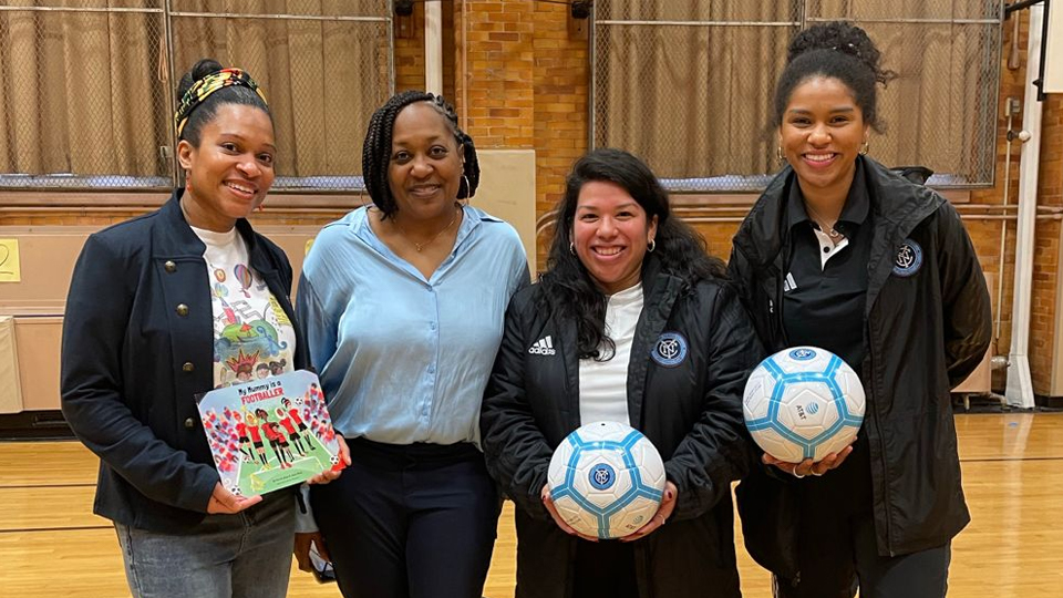 Four people stand in a sports hall. Kerrine Bryan is pictured on the left holding a copy of her book. The two people on the right of the image each hold a football.