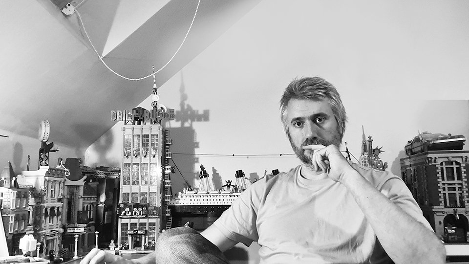 A black and white image of Pete Wharmby. He is seated in a room with model buildings and a ship around him.  