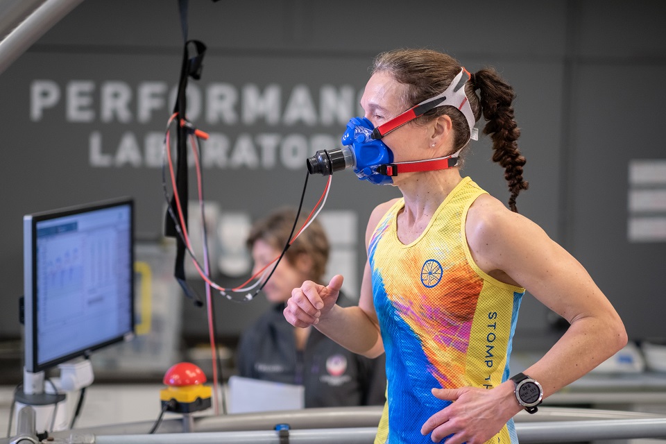 Someone running indoors wearing a mask device. There is a person out of focus in the background. There is a computer screen on the left of the image.