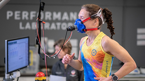 Someone running indoors wearing a mask device. There is a person out of focus in the background. There is a computer screen on the left of the image.