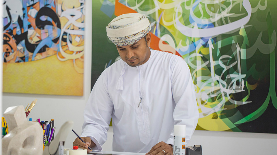 Alumnus Salman AlHajri stood at a desk drawing with artwork on the walls behind them.