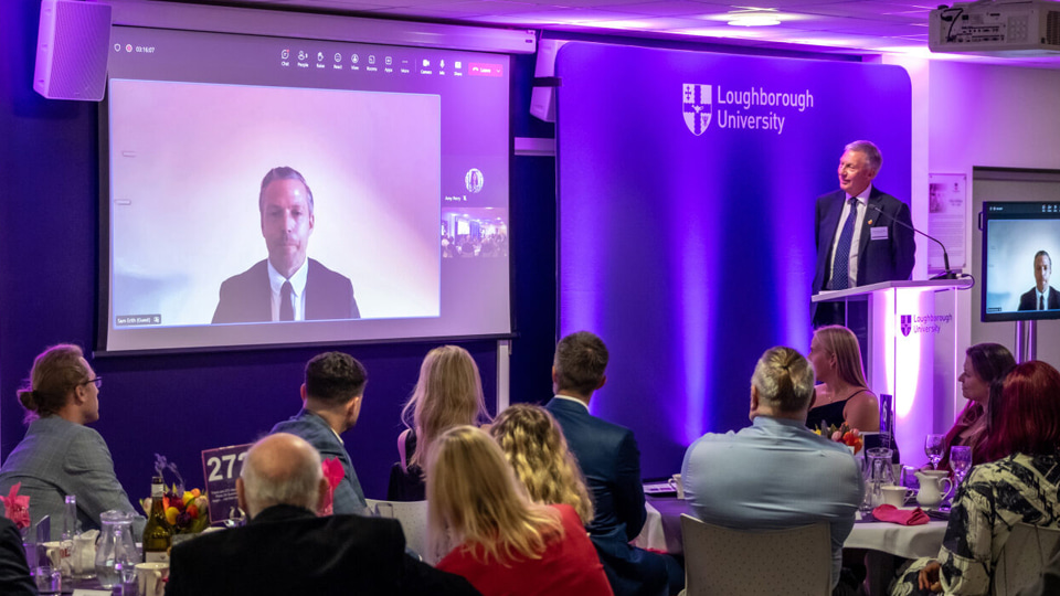 David Moorcroft looks to a screen to video interview Sam Erith. There are people seated at tables looking towards the screen.