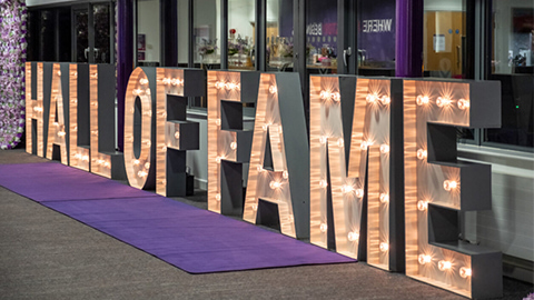 Large lit-up letters spelling out Hall of Fame, standing behind a purple carpet and in front of a set of windows