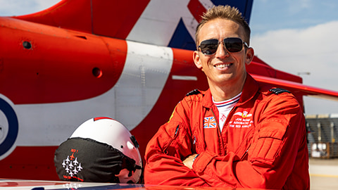 Jon Bond stands arms folded leaning on the wing of a plane. He is wearing a red flying suit and sunglasses. To the left is his helmet and in the background is more of the Red Arrows plane.