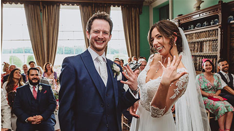 Lizzie and Lewis on their wedding day with guests in the background and Lizzie holding up her hand to show her ring.