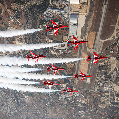 Eight Red Arrows planes flying over Malta. Smoke is coming from the back of each plane, with the view of the ground beneath them.
