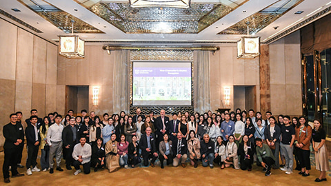 A photo of a large group of people standing and crouching together for the photo. There is a screen on the wall behind them with a photo of campus on. There is a high ceiling and big square lights hanging down.