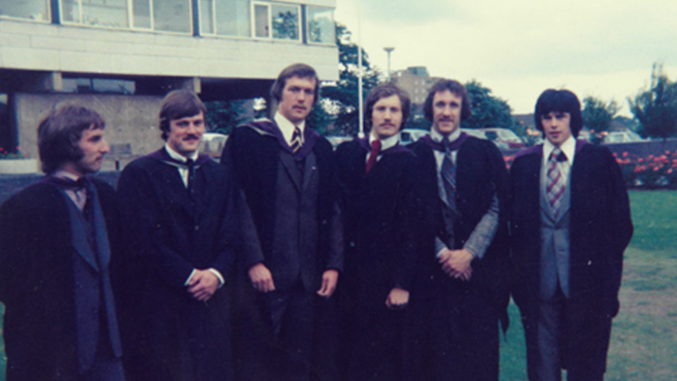 A graduation photo taken in 1974. The group of people stand on grass in front of a building. They are wearing gowns.