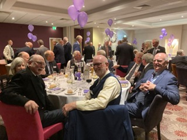 People seated at a table wearing smart clothes. Balloons are on the tables. There are people standing in the background.