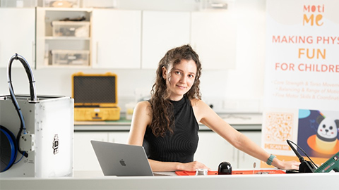 Katie seated at a desk with a laptop. There are cupboards on the wall behind her. On the desk is a piece of machinery.