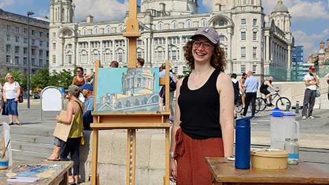 Monica stands next to an easel with her painting on. There is a building in the background.