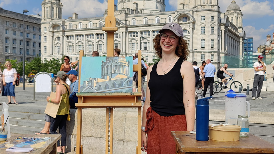 Monica stands next to an easel with her painting on. There is a building in the background.