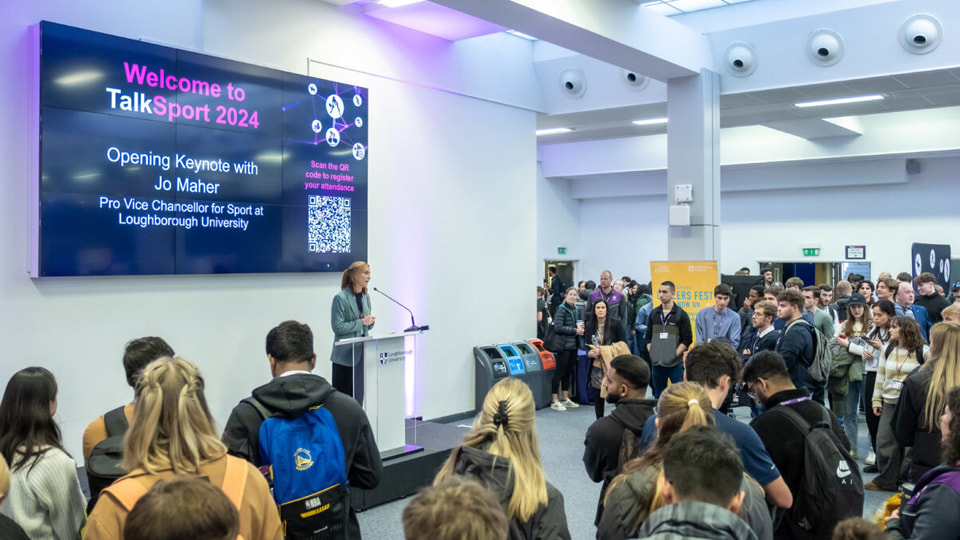 People standing looking towards someone on a stage, speaking. There is a screen on the wall behind the stage with “Welcome to TalkSport 2024” on.