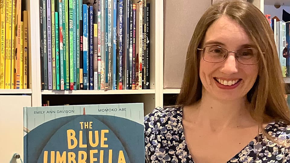 Emily is holding up her new book 'The Blue Umbrella' in front of a shelf of books and she is smiling.