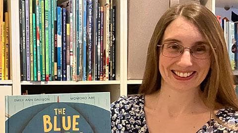 Emily is holding up her new book 'The Blue Umbrella' in front of a shelf of books and she is smiling.