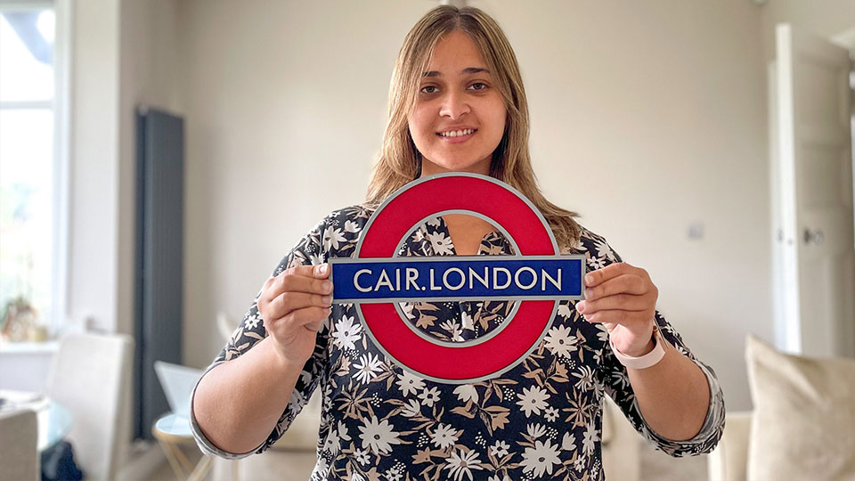 Tanya is smiling and holding up a London Underground logo sign but the text says 'CAIR.London'