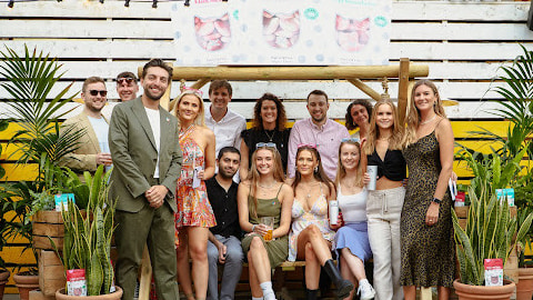 A group photo of people taken outdoors. There's a few plants. People are dressed in summery clothes.