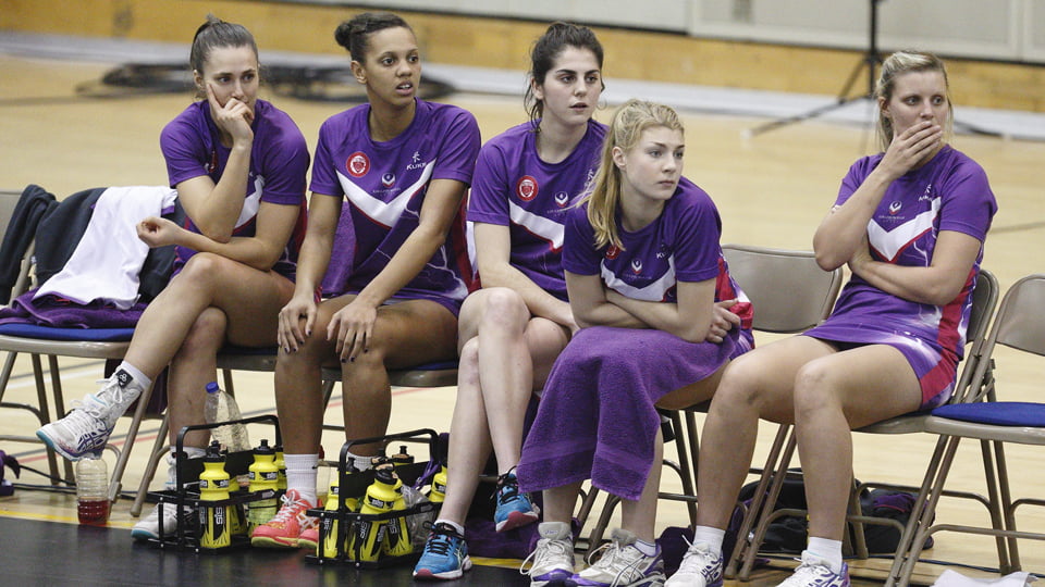 A group of five people seated on chairs in a sports hall wearing purple netball kit. They have concerned looks on their faces.