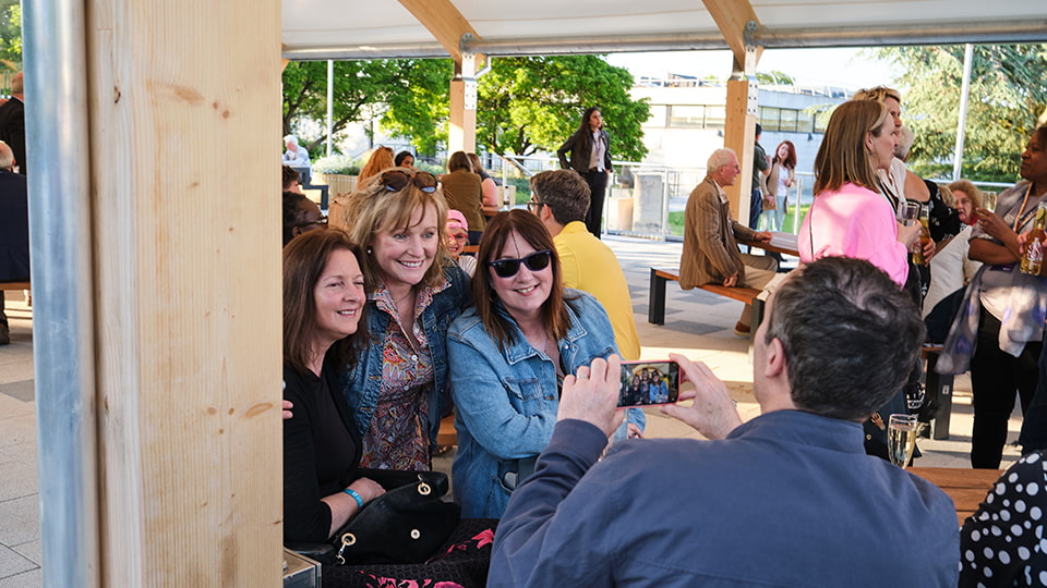 Three people having a photo taken by another person. There are other people in the background.