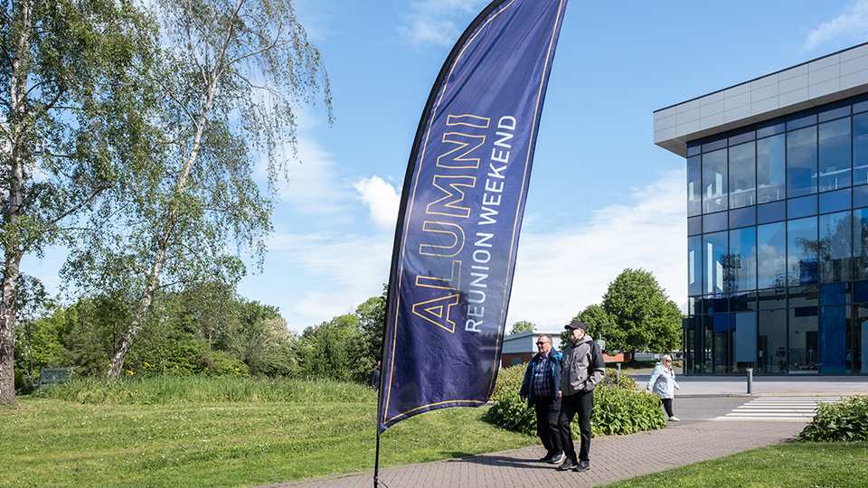 A purple flag on a stick with text, reads: Alumni Reunion Weekend. Behind the flag is a glass fronted building. There is grass and trees.
