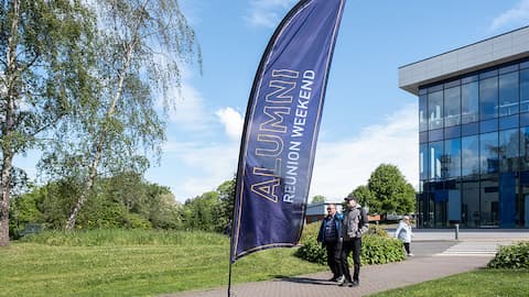 A purple flag on a stick with text, reads: Alumni Reunion Weekend. Behind the flag is a glass fronted building. There is grass and trees.