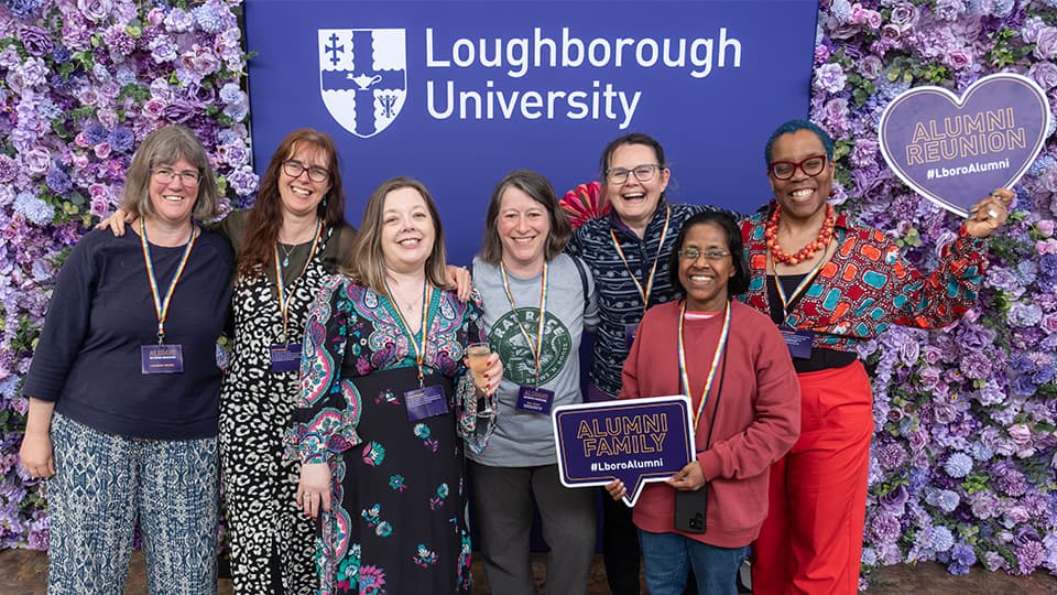 A group of people stand together smiling. Behind them is a purple and flower backdrop. A couple of people hold signs that say Alumni Reunion and Alumni Family.