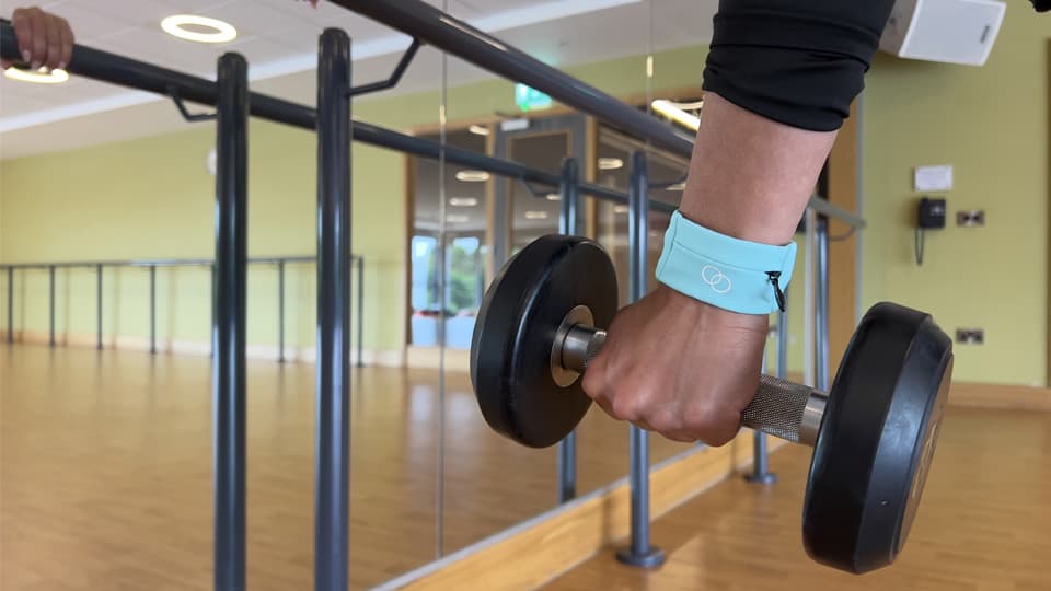Someone's hand and arm. They are holding a dumb bell and wearing a Ring Buddy wristband. There is a mirror showing the reflection.