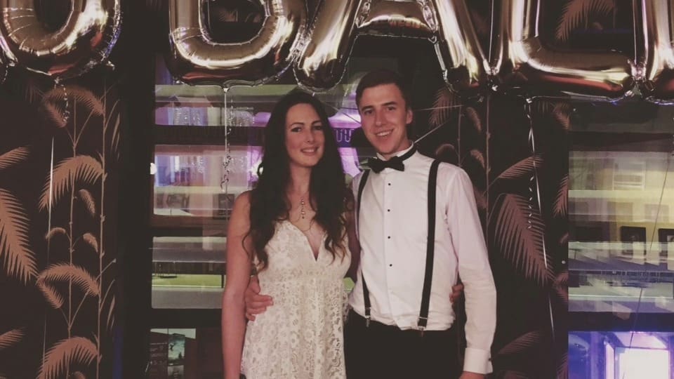 Two people standing smiling in evening wear. There are balloons above their heads.