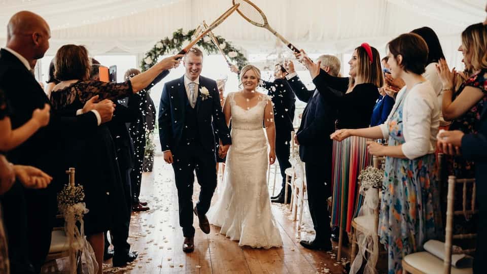 A couple wearing wedding attire walking down an aisle. There are tennis rackets held above their heads by guests who are standing each side of the aisle.
